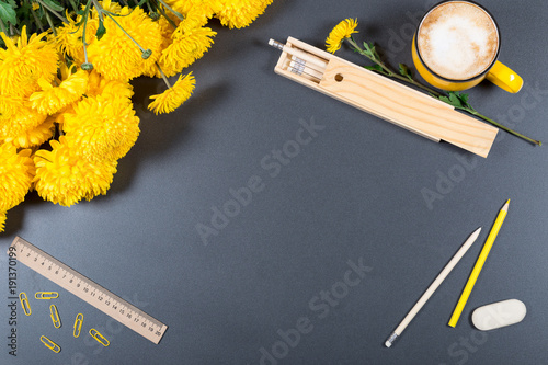Gray desk surface with color pencils, eraser, ruller, wooden pencil box, big cup of cappuccino and bunch of yellow chrysanthemums. Mock-up. Top view. Flat lay.