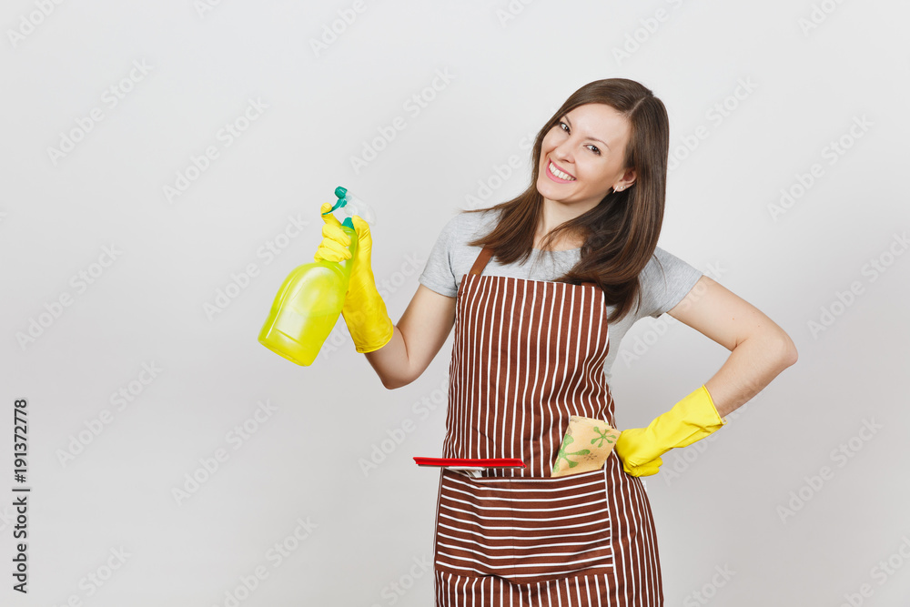 Young smiling housewife in yellow gloves, striped apron, cleaning rag, squeegee in pocket isolated on white background. Housekeeper woman holding spray bottle with cleaner liquid. Bottle copy space. Young smiling housewife in yellow gloves, striped apron, cleaning rag, squeegee in pocket isolated on white background. Housekeeper woman holding spray bottle with cleaner liquid. Bottle copy space.