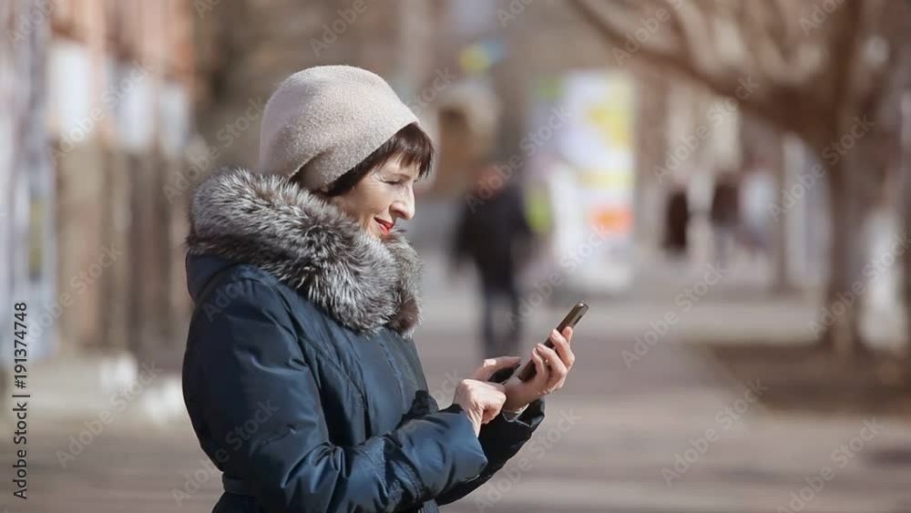 An impressive view of a middle-aged woman who stands in profile in a picturesque alley and touches the screen of her smartphone on a sunny day An impressive view of a middle-aged woman who stands in profile in a picturesque alley and touches the screen of her smartphone on a sunny day