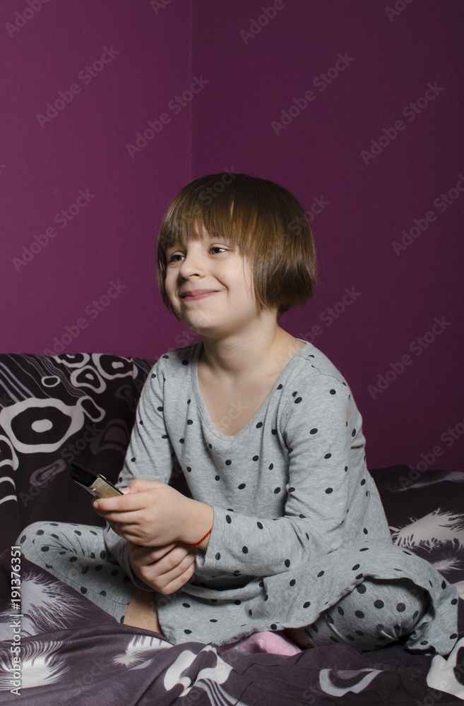 A small, beautiful blue-eyed girl smiles and watches TV, holding a remote control A small, beautiful blue-eyed girl smiles and watches TV, holding a remote control