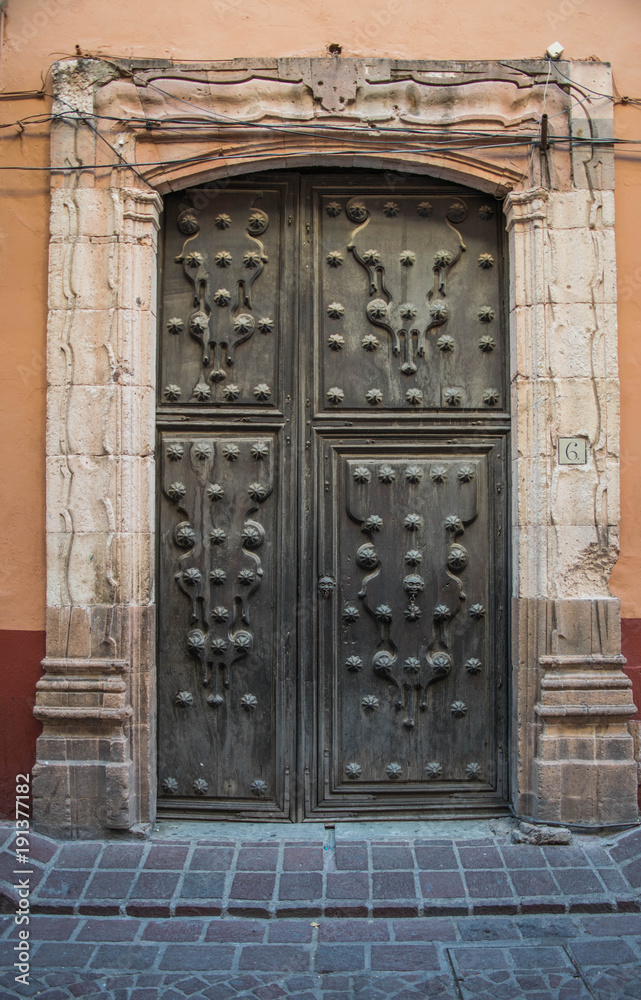 An old elaborate door, with beautifully crafted details and a massive stone frame, with a brick walkway, in Guanajuato, Mexico