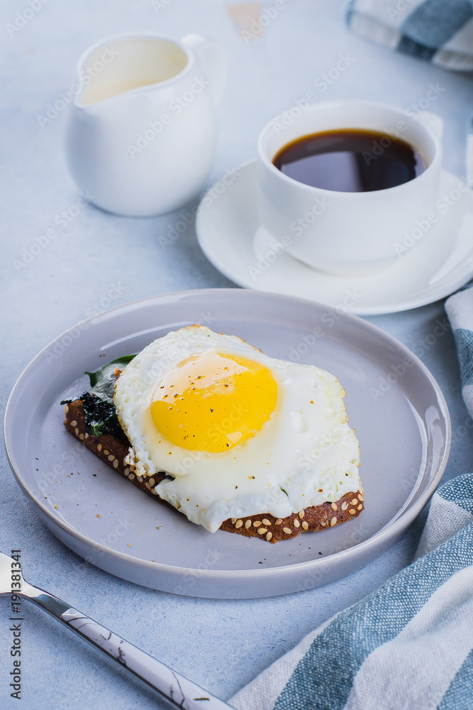 Rye bread toasts with fried spinach and egg with cup of coffee and milk on blue table background. Healthy Breakfast Food Concept. Rye bread toasts with fried spinach and egg with cup of coffee and milk on blue table background. Healthy Breakfast Food Concept.