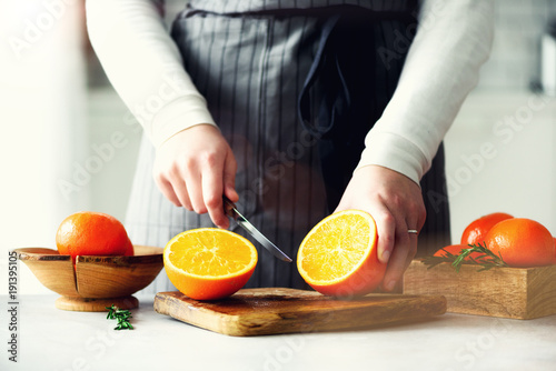 Woman hands slicing orange, cutting citrus fruit. Knife, wooden cutting board on design white kitchen. Copy space