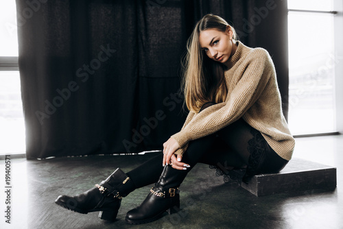Amazing young girl in stylish clothes is posing sitting on the floor indoors near a black background