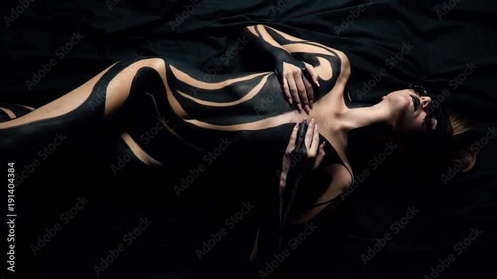 A girl in a lace mask and black body art lies on a black sheet