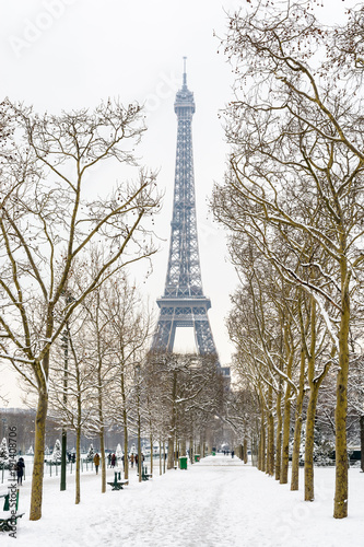Winter in Paris in the snow. The Eiffel tower seen from the Champ de Mars with a snow covered tree lined alley in the foreground.