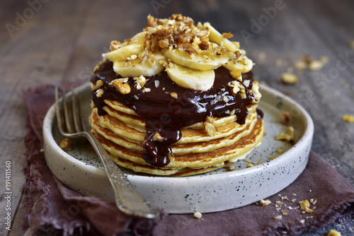 Stack of homemade delicious banana pancakes topped with chocolate sauce, banana slices and walnut.