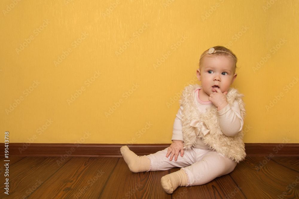  little girl with a finger in mouth dressed in beautiful clothes resting sits on a wooden floor in the children's room.baby on yellow background. place for text.
