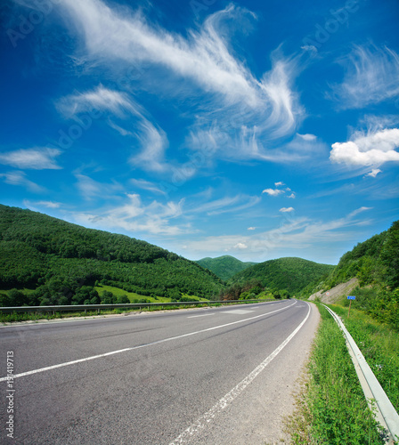 Asphalt road in summer green forest mountains on blue sky background
