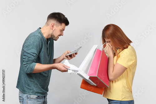 Angry man scolding his wife who has spent a lot of money for shopping, against light background