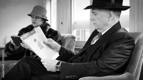 Travelers on a passenger train dressed in stylish 1940s period attire sitting in the lounge car of a vintage railcar with the man reading a newspaper article and the woman a book.