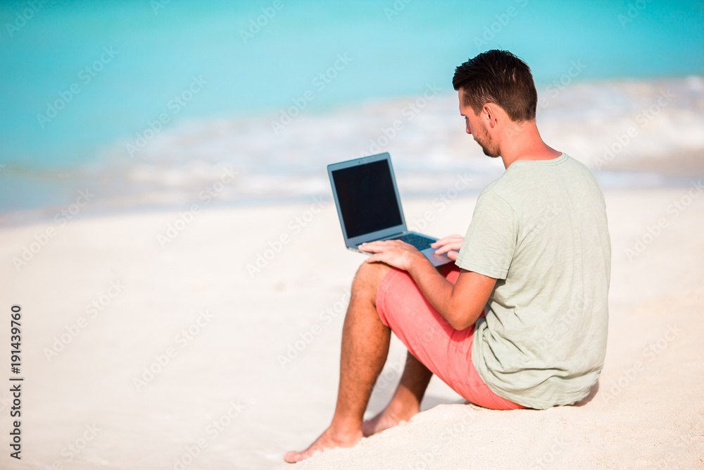 Young man sitting on sand with laptop on tropical caribbean beach. Man with computer and working on the beach