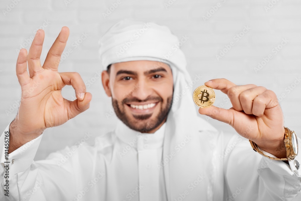 Studio portrait of a smiling Arabian man keeps bitcoin and looks very happy, standing on white background. He wears traditional white Arabian costume,golden watch and cufflinks. Studio portrait of a smiling Arabian man keeps bitcoin and looks very happy, standing on white background. He wears traditional white Arabian costume,golden watch and cufflinks.