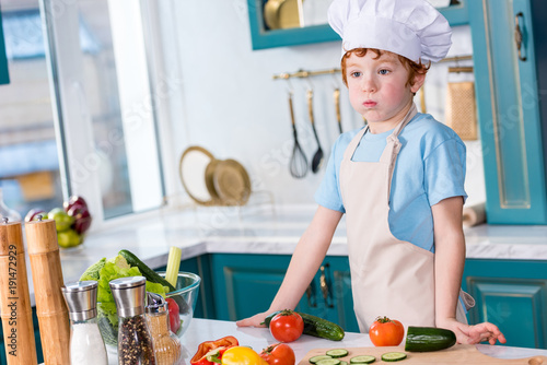 cute little boy in chef hat and apron looking away while cooking in kitchen