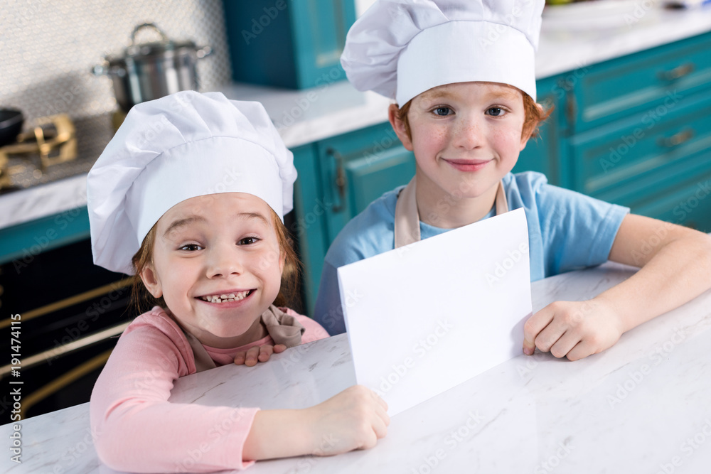 cute little children in chef hats holding blank card and smiling at camera cute little children in chef hats holding blank card and smiling at camera