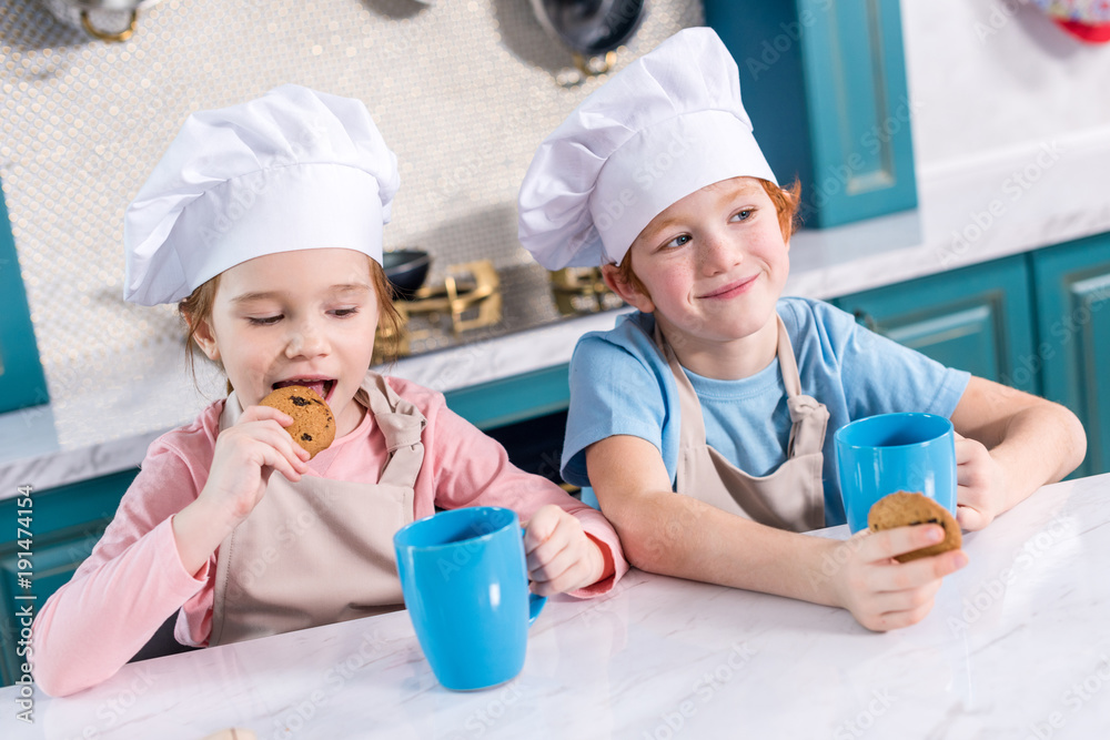 smiling children in chef hats drinking tea and eating cookies in kitchen smiling children in chef hats drinking tea and eating cookies in kitchen