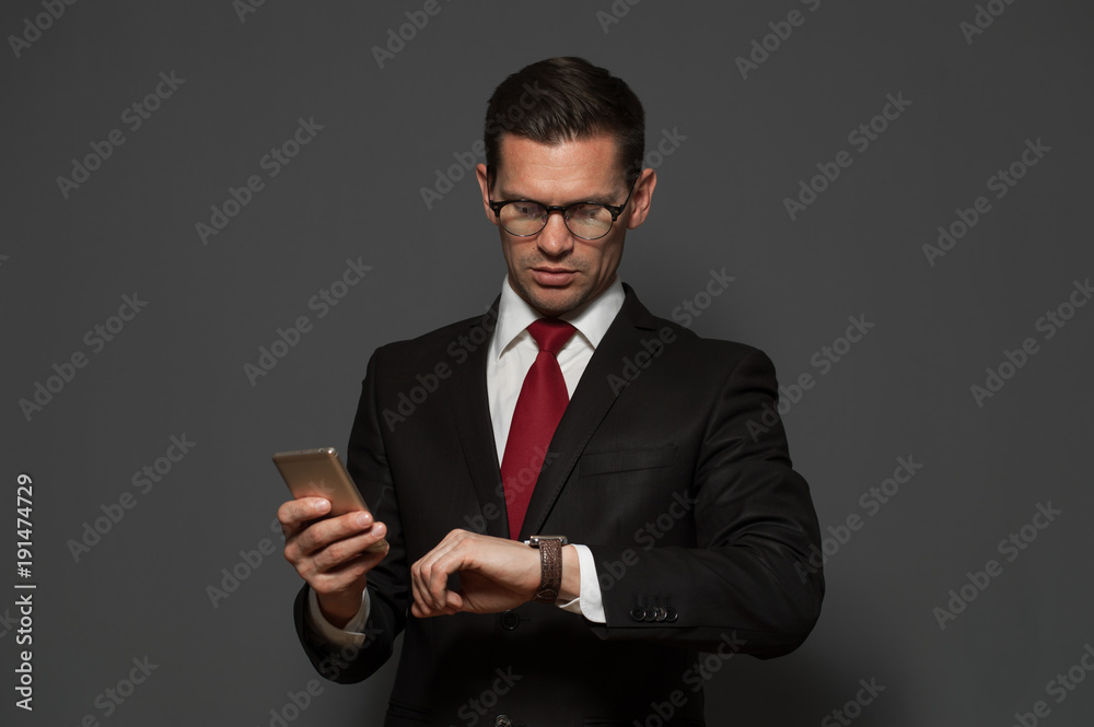 Successful young man hurrying on meeting and looking at his watch with smartphone in his hand on gray background. Successful young man hurrying on meeting and looking at his watch with smartphone in his hand on gray background.