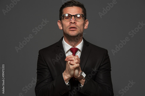 Portrait of middle-aged businessman in formal suit and glasses with crossed hand near his chest and praying against gray background