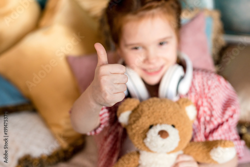 cute little child in headphones holding teddy bear and showing thumb up