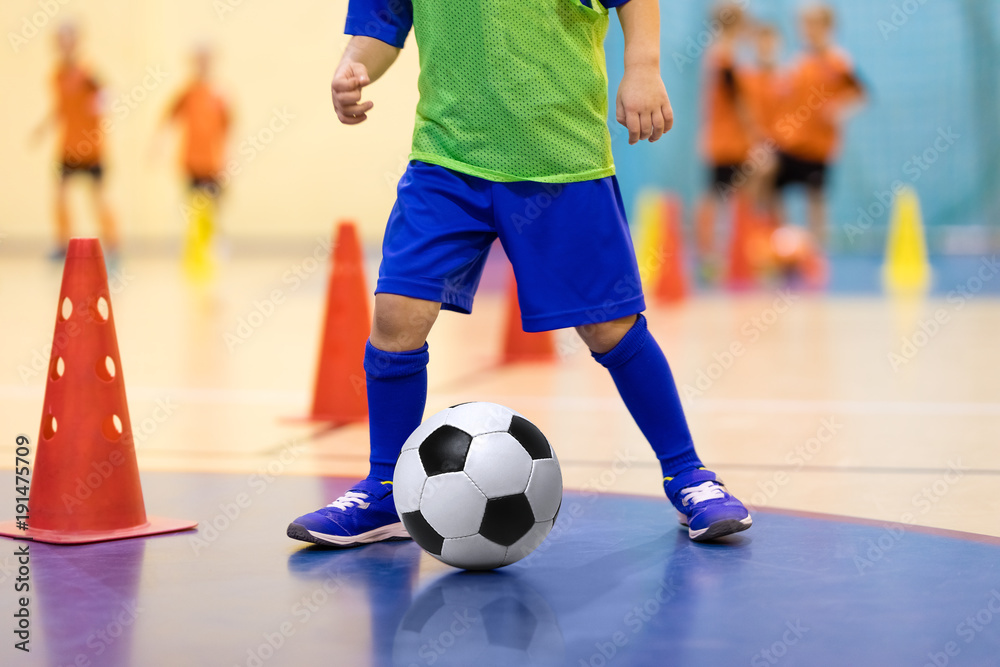 Naklejka premium Football futsal training for children. Soccer training dribbling cone drill. Indoor soccer young player with a soccer ball in a sports hall. Player in blue uniform. Sport background.