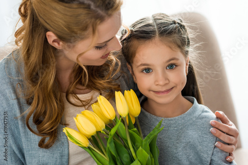 happy mother looking at cute daughter with yellow tulips smiling at camera