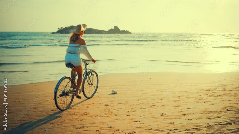 Woman in white and hat mooving on her bicycle along beach sand at summer time under burning sunset sky, lifestyle carefree. Woman in white and hat mooving on her bicycle along beach sand at summer time under burning sunset sky, lifestyle carefree.