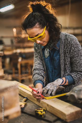 Close up view of hardworking focused professional serious carpenter woman holding ruler and pencil while making marks on the wood at the table in the fabric workshop.