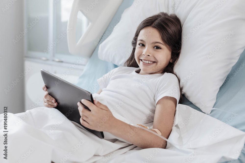 No time for worries. Adorable child grinning broadly while lying in a hospital bed with a drop counter in her arm and staying positive during her treatment. No time for worries. Adorable child grinning broadly while lying in a hospital bed with a drop counter in her arm and staying positive during her treatment.