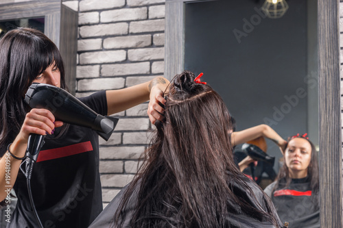 Beautiful hair stylist with tattooed hands using a hair dryer on hair of young brunette woman