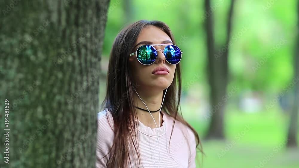 Young woman stands near tree in city park listens to music and dances. Beautiful girl in sunglasses with earpieces listens to music on the nature Young woman stands near tree in city park listens to music and dances. Beautiful girl in sunglasses with earpieces listens to music on the nature