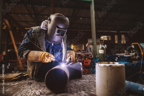 Welder in protective uniform and mask welding metal pipe on the industrial table while sparks flying.