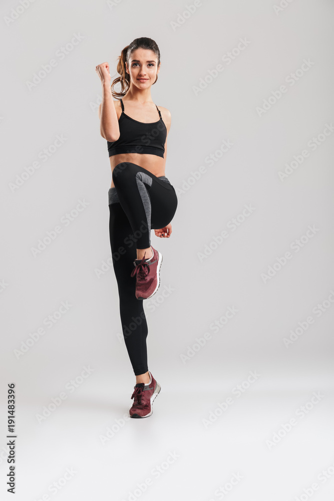 Full-length portrait of attractive pretty female trainer of instructor in sportswear warming up and doing exercises, isolated against gray background