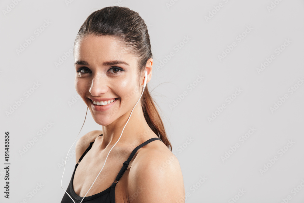 Portrait closeup of young smiling woman in gym listening to music using earphones and smartphone, isolated over gray background Portrait closeup of young smiling woman in gym listening to music using earphones and smartphone, isolated over gray background