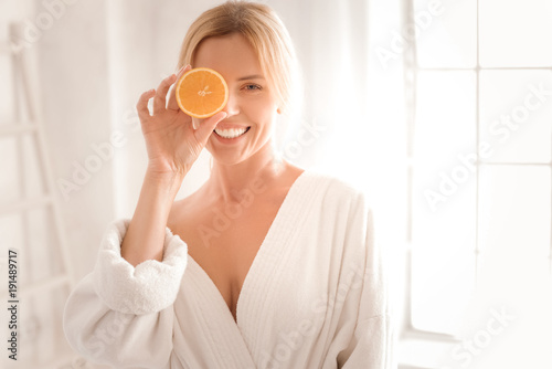 Fresh energy. Happy delighted nice woman holding an orange half and smiling while using it for cosmetic procedures
