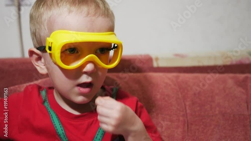 A child in safety glasses sits on the sofa at home and watches TV. Eat corn sticks and smile