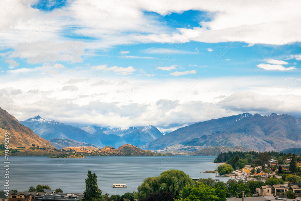 View of Wanaka lake and alpine resort town with the mountain range in the background in New Zealand, on a cloudy summer day. 
