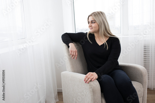  white blonde girl sitting on a white armchair on a white background in black pullover and posing and smiling with her smartphone talking with friends