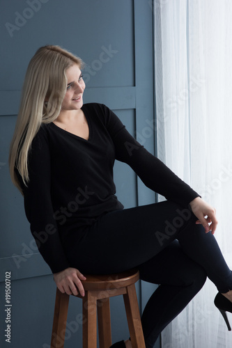 white blonde girl sitting on a high bar chair on a grey dark grey background in black pullover and black high heels with red basis posing and smiling 