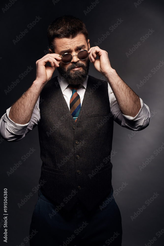 Obraz premium Low key portrait of serious man with dark hair, mustache and beard in white shirt, brown vest and colorful tie takes off round sunglasses