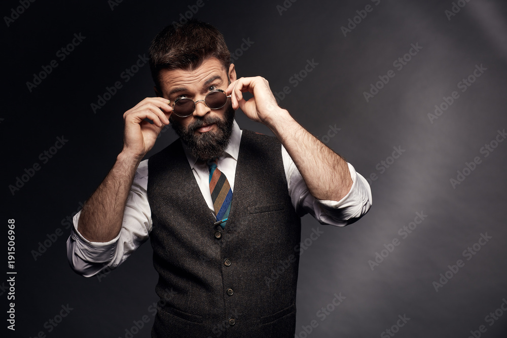 Studio portrait of attractive handsome man with dark hair, mustache and beard in white shirt, brown vest and colorful tie takes off round sunglasses Studio portrait of attractive handsome man with dark hair, mustache and beard in white shirt, brown vest and colorful tie takes off round sunglasses