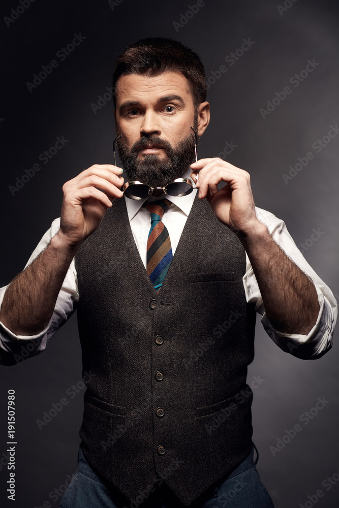 Studio portrait of attractive handsome man with dark hair, mustache and beard in white shirt, brown vest, colorful tie with round sunglasses in hands Studio portrait of attractive handsome man with dark hair, mustache and beard in white shirt, brown vest, colorful tie with round sunglasses in hands