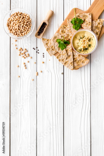Serve hummus. Bowl with dish near pieces of crispbread on white wooden background top view copy space