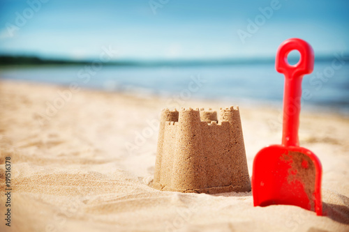 Fototapeta Naklejka Na Ścianę i Meble -  Sandcastle with a shovel on the sea in summertime. Seashore on beautiful day. Sand on the beach and blue water