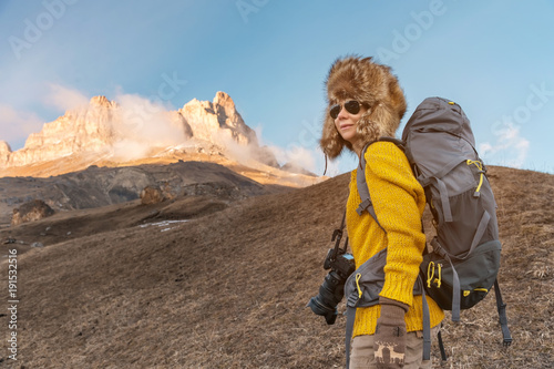 The backpacker girl in sunglasses and a big northern fur hat with a backpack on her back is standing on a rock and looking toward the cliffs hiding in the rock.