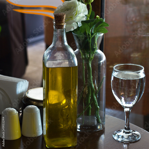 A bottle of olive oil on the table. Two glasses of water. Vase with Flowers. The salt cellar