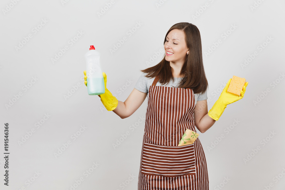 Young housewife in yellow gloves, striped apron, cleaning rag in pocket isolated on white background. Woman holding bottle with cleaner liquid for washing dishes, sponge. Copy space for advertisement. Young housewife in yellow gloves, striped apron, cleaning rag in pocket isolated on white background. Woman holding bottle with cleaner liquid for washing dishes, sponge. Copy space for advertisement.