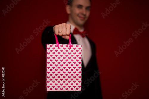 Young smiling man in suit hold love gift bag, with red background, focus on gift bag. Valentines day composition