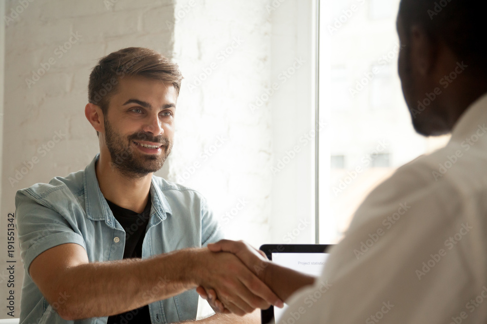 Fototapeta premium Smiling confident caucasian candidate handshaking african hr manager at job interview, white man client or customer greeting black service supplier, multi-ethnic businessmen shaking hands at meeting