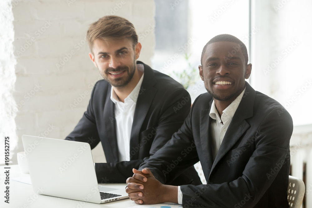 Multiracial business professionals team portrait, smiling african and caucasian businessmen in suits sitting at office desk with laptop, black and white successful startup founders looking at camera Multiracial business professionals team portrait, smiling african and caucasian businessmen in suits sitting at office desk with laptop, black and white successful startup founders looking at camera