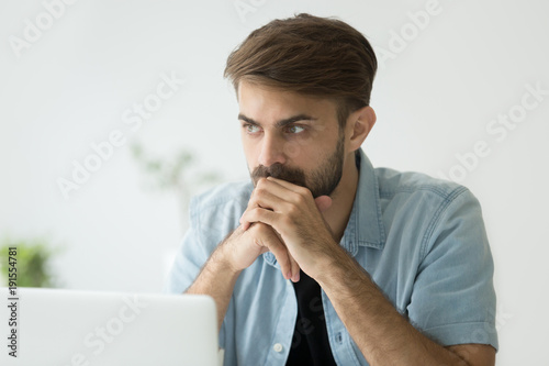 Thoughtful serious young man lost in thoughts in front of laptop, focused businessman or absent-minded student thinking of problem solution, worried puzzled manager pondering question at work photo